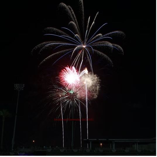 Large red and white peony fireworks shell breaking above a stadium with a blue palm-tree burst rising beside it at night
