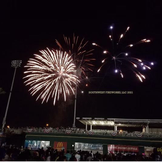 Two large fireworks shells breaking above a lit baseball stadium filled with spectators after a game