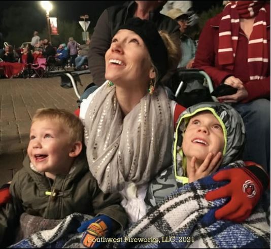 Mother and two young sons wrapped in a blanket smiling and looking up at a fireworks display on a cool evening in a crowd of seated spectators