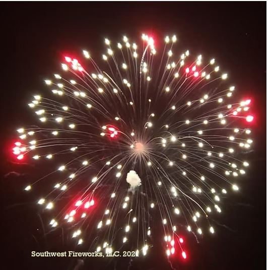 Large symmetrical fireworks shell break with white radial trails and bright red pistil dots filling the night sky