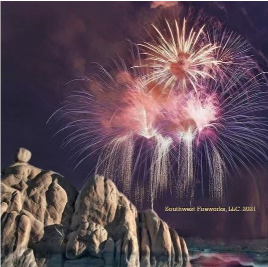 Pink and gold fireworks shells breaking above a rugged rock outcropping and reservoir at night