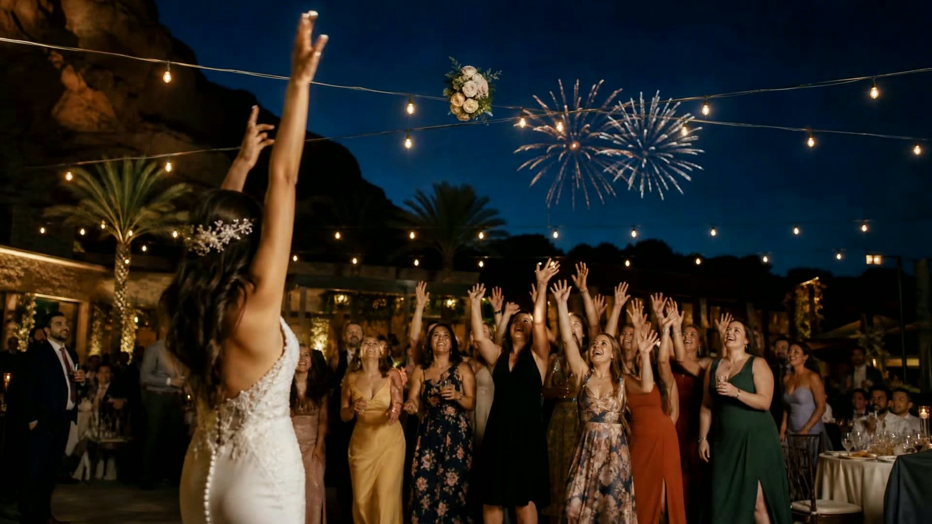 A bride tossing her bouquet backward toward laughing wedding guests as professional aerial fireworks bloom in the distant Arizona night sky