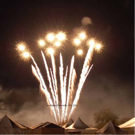Bright white comet trails rising into a row of gold shell breaks above event tents during the Skies Over Bagdad fireworks finale