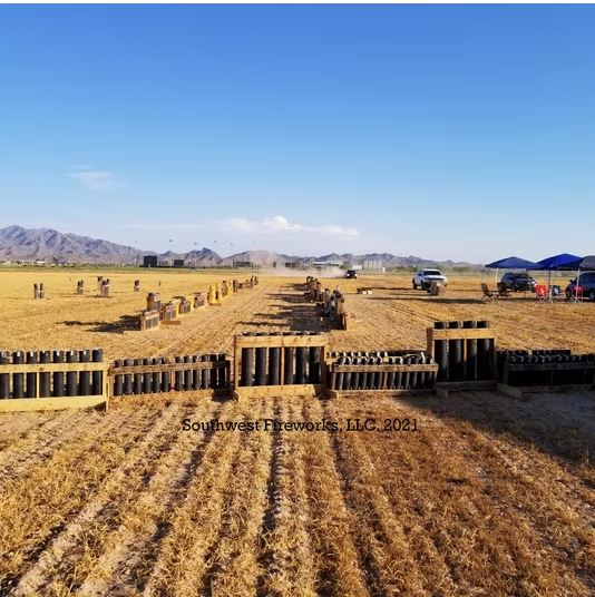Long rows of loaded fireworks mortar racks staged across a wide open Arizona field with mountains and trucks in the background at daytime