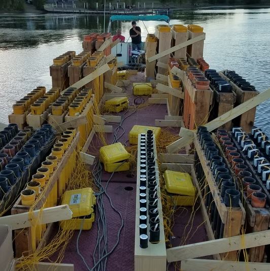 Fireworks barge on water loaded with rows of mortar tubes, electric firing modules, and yellow shot boxes at dusk before a waterfront display