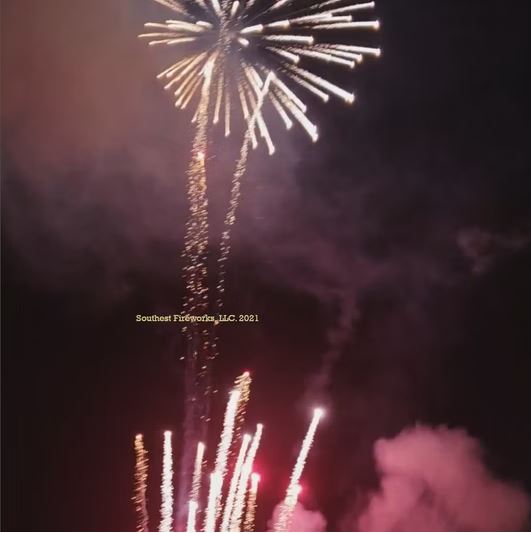 Opening shell of a fireworks finale — a single bright gold burst high in the sky with rising comet tails and pink smoke drifting below