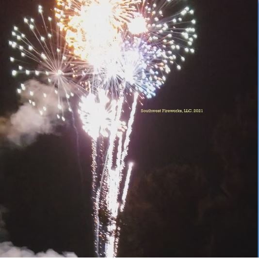 Multiple large white and gold fireworks shells breaking simultaneously above a tree line during a professional show finale