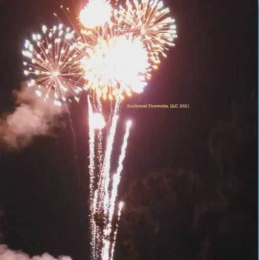 Three large chrysanthemum fireworks shells breaking together above a tree line with bright ascending comet trails