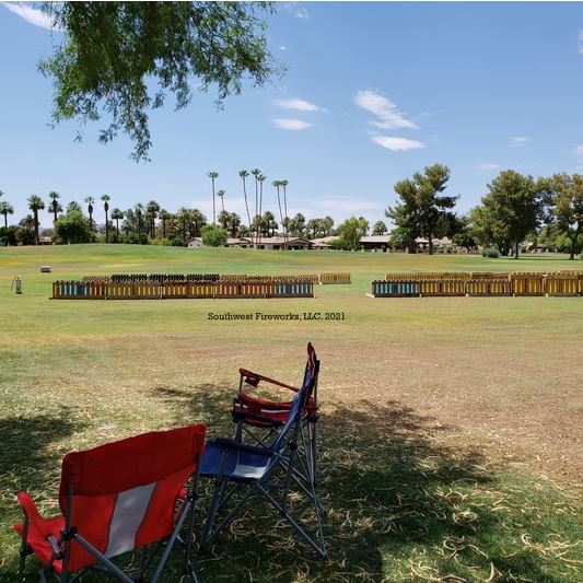 Two empty red and blue lawn chairs facing a long line of loaded fireworks mortar racks on a green golf course with palm trees and blue sky before showtime