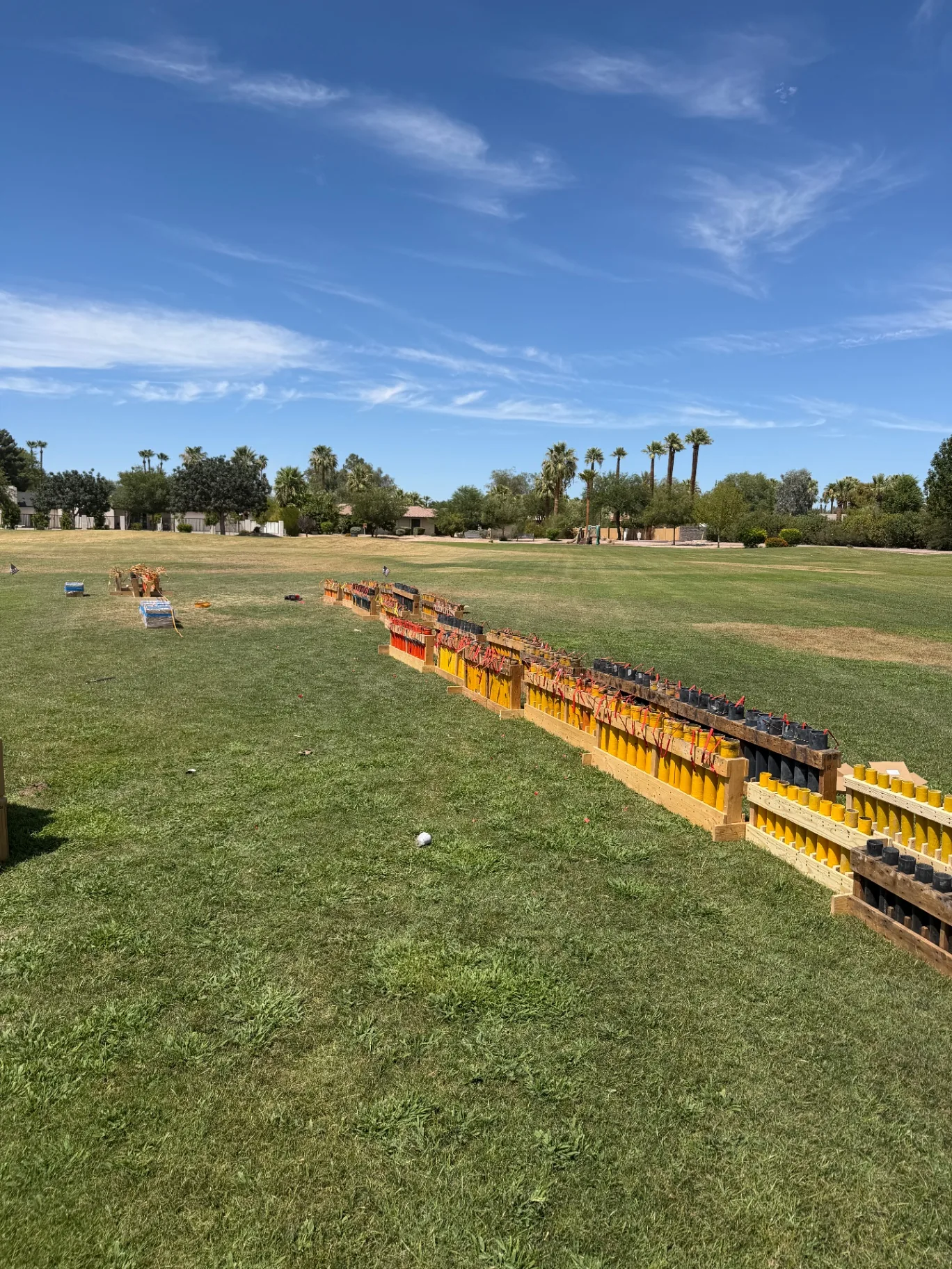 Row of loaded yellow and black fireworks mortar tubes secured in wooden racks on a grass field with palm trees in the background