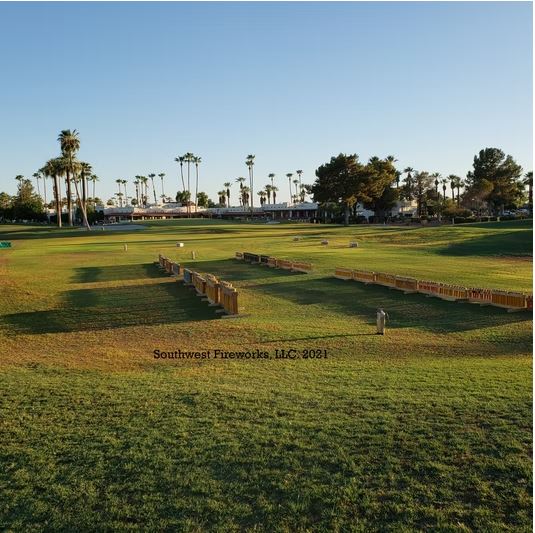 Long rows of loaded fireworks mortar racks staged across a manicured Arizona golf course fairway with palm trees and a clubhouse in the background at golden hour