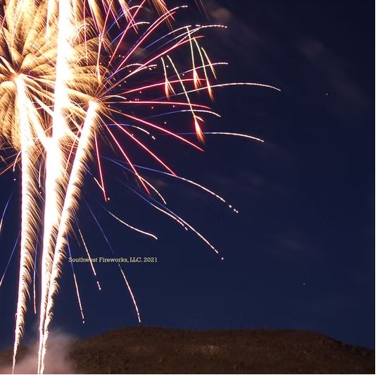 Long ascending gold comet trails leading up to a large red and blue aerial shell break above a dark mountain silhouette