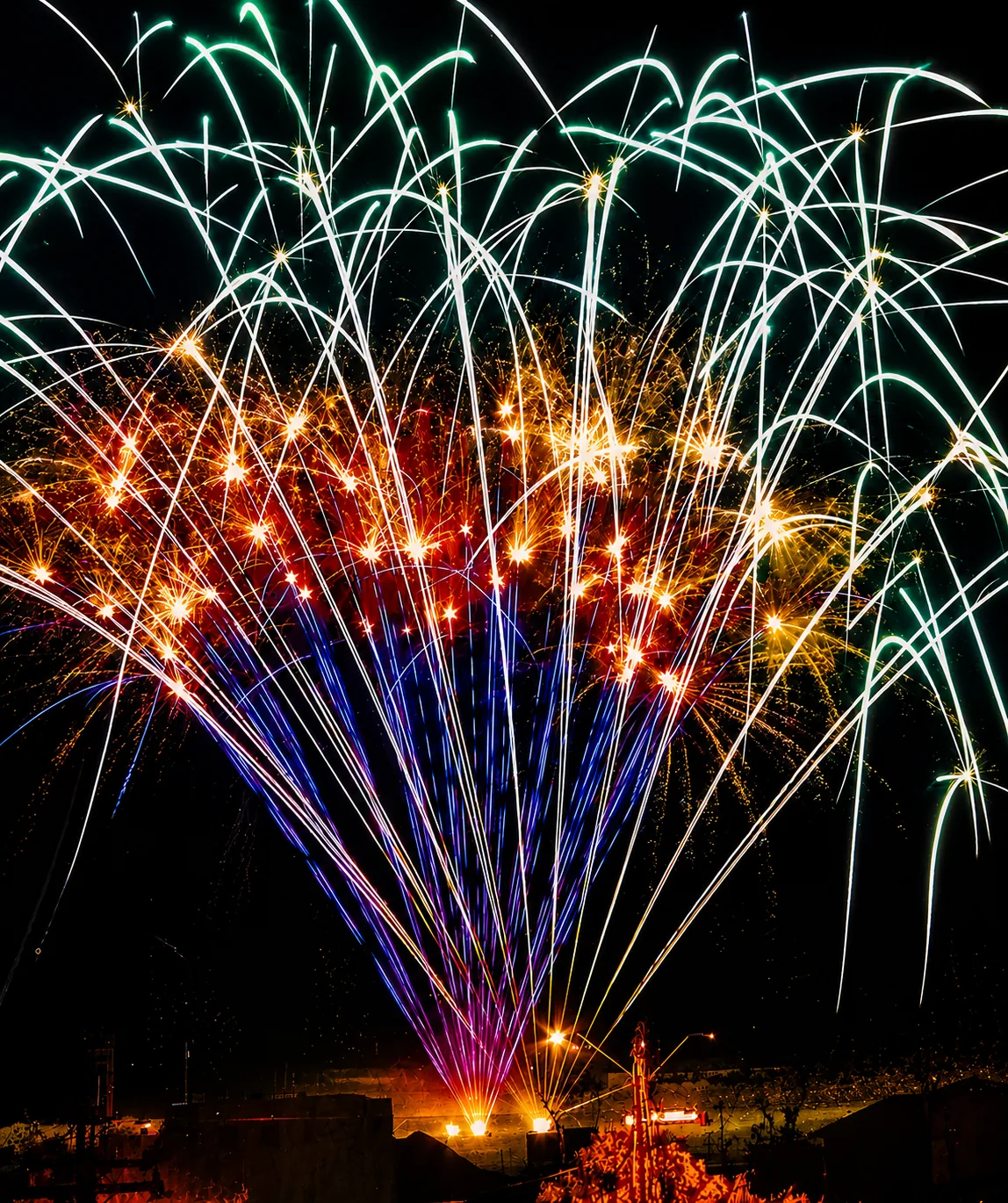 Multi-shot fireworks cake firing a rapid sequence of green, gold, blue, and red aerial bursts across the night sky
